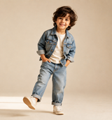 A young child wearing a light blue denim jacket and matching denim jeans over a cream t-shirt, paired with beige sneakers, posing against a warm beige studio backdrop.