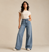 A woman modeling a pair of high-waisted, wide-leg light blue denim jeans with a white t-shirt, standing against a plain beige studio backdrop.
